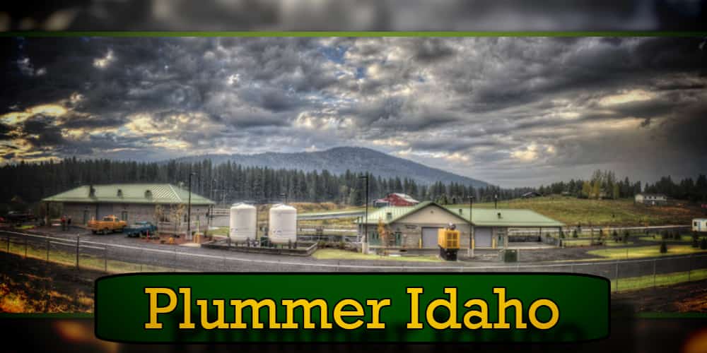 A small industrial area in Plummer, Idaho with buildings, storage tanks, and pine trees. A cloudy sky and hills in the background. A tow truck is parked near one of the buildings. Text at the bottom reads