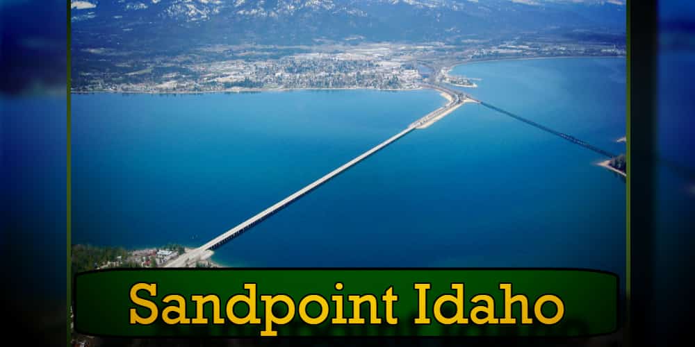 Aerial view of Sandpoint, Idaho, featuring a long bridge over Lake Pend Oreille with mountains in the background. A tow truck is crossing the bridge. The text 
