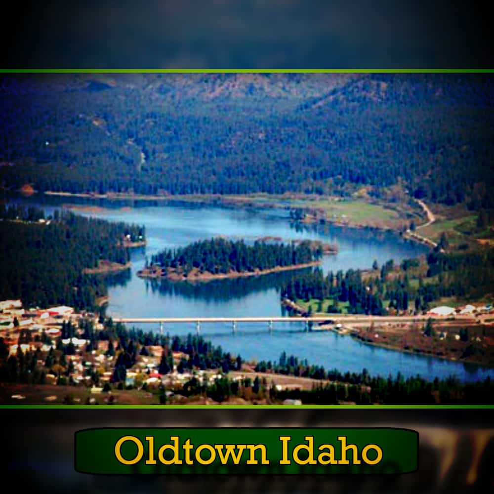 Aerial view of Oldtown, Idaho, showing a lake surrounded by forested hills and a bridge connecting parts of the town, with a tow truck making its way across the bridge.
