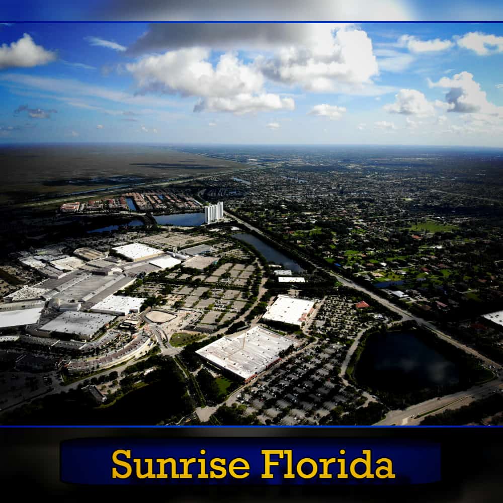 Aerial view of Sunrise, Florida, showing commercial buildings, residential areas, and a network of roads and waterways under a partly cloudy sky with tow trucks navigating the streets.