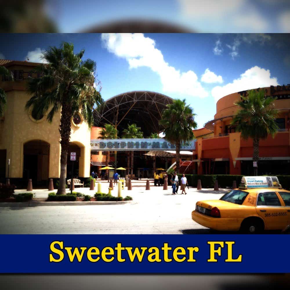Outdoor view of a plaza in Sweetwater, FL with palm trees, a yellow taxi, and a glass canopy entrance. Buildings with yellow and orange facades line the space under a clear sky, with a tow truck idling nearby.
