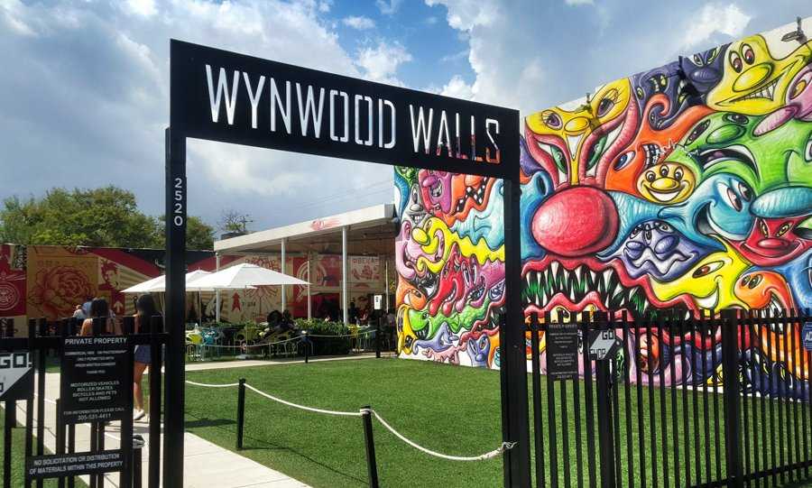 Entrance to Wynwood Walls, showcasing vibrant graffiti art on walls against a partly cloudy sky. Visitors can be seen in the background near an outdoor seating area, while a nearby tow truck subtly reminds us of the bustling city life.