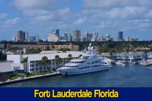 A yacht is docked near a marina with buildings and skyscrapers in the background under a partly cloudy sky. A tow service boat stands ready nearby. Text at the bottom reads: 
