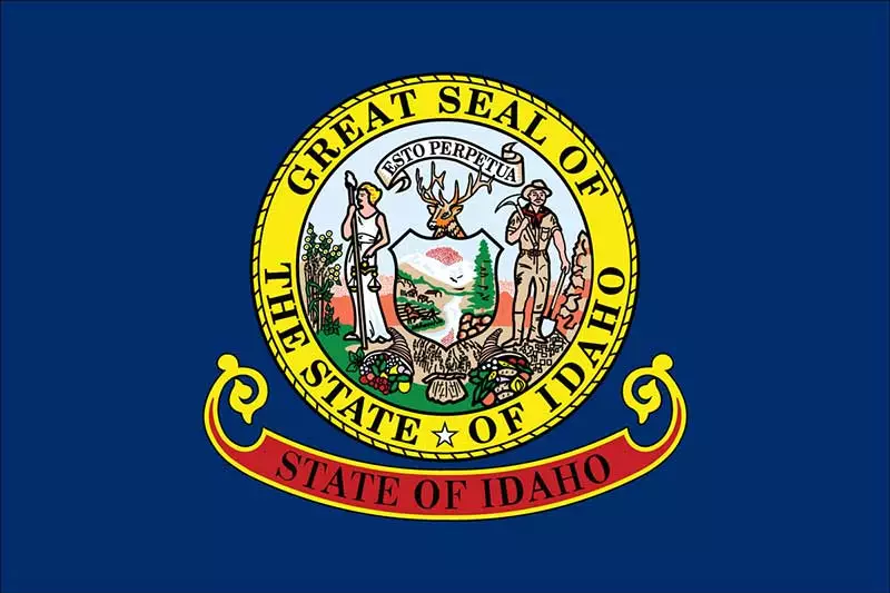 The image shows the state flag of Idaho, featuring the Great Seal of Idaho in the center, surrounded by a yellow circle on a blue background, with 