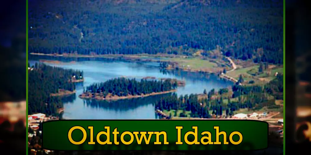 Aerial view of Oldtown, Idaho featuring a lake surrounded by forested land and scattered buildings, with a tow truck parked near the lake. The text 