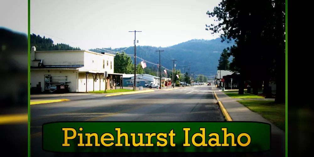 A quiet street in Pinehurst, Idaho, featuring small buildings on either side, with mountains in the background and a large sign in the foreground displaying the town's name, while a tow truck idles nearby.