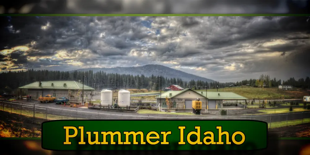 A small industrial area in Plummer, Idaho with buildings, storage tanks, and pine trees. A cloudy sky and hills in the background. A tow truck is parked near one of the buildings. Text at the bottom reads 