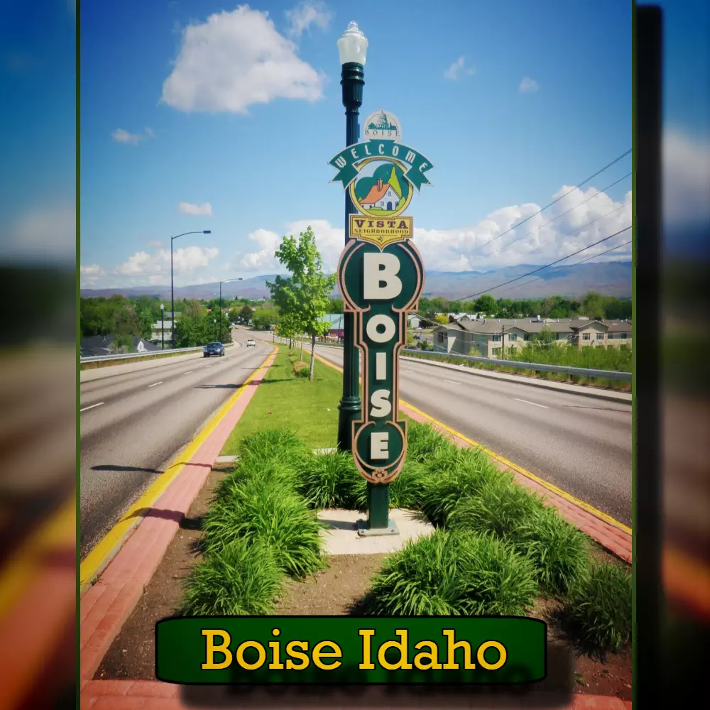 Roadside sign in Boise, Idaho, featuring the city name and welcoming visitors. The sign is surrounded by greenery, with a view of a street lined with occasional tow trucks and distant hills in the background.