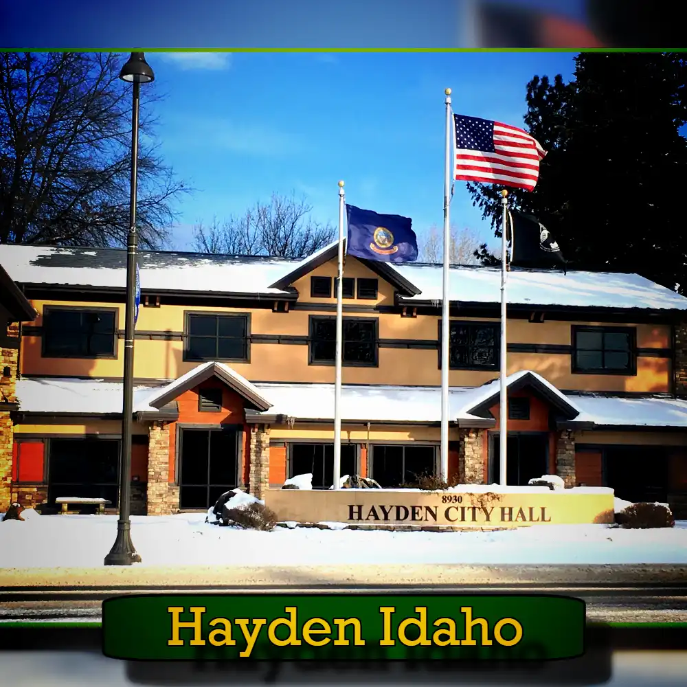 Exterior view of Hayden City Hall in Hayden, Idaho, with the American and Idaho state flags flying in front. The building is surrounded by snow, and a tow truck is parked nearby, adding to the picturesque winter scene.
