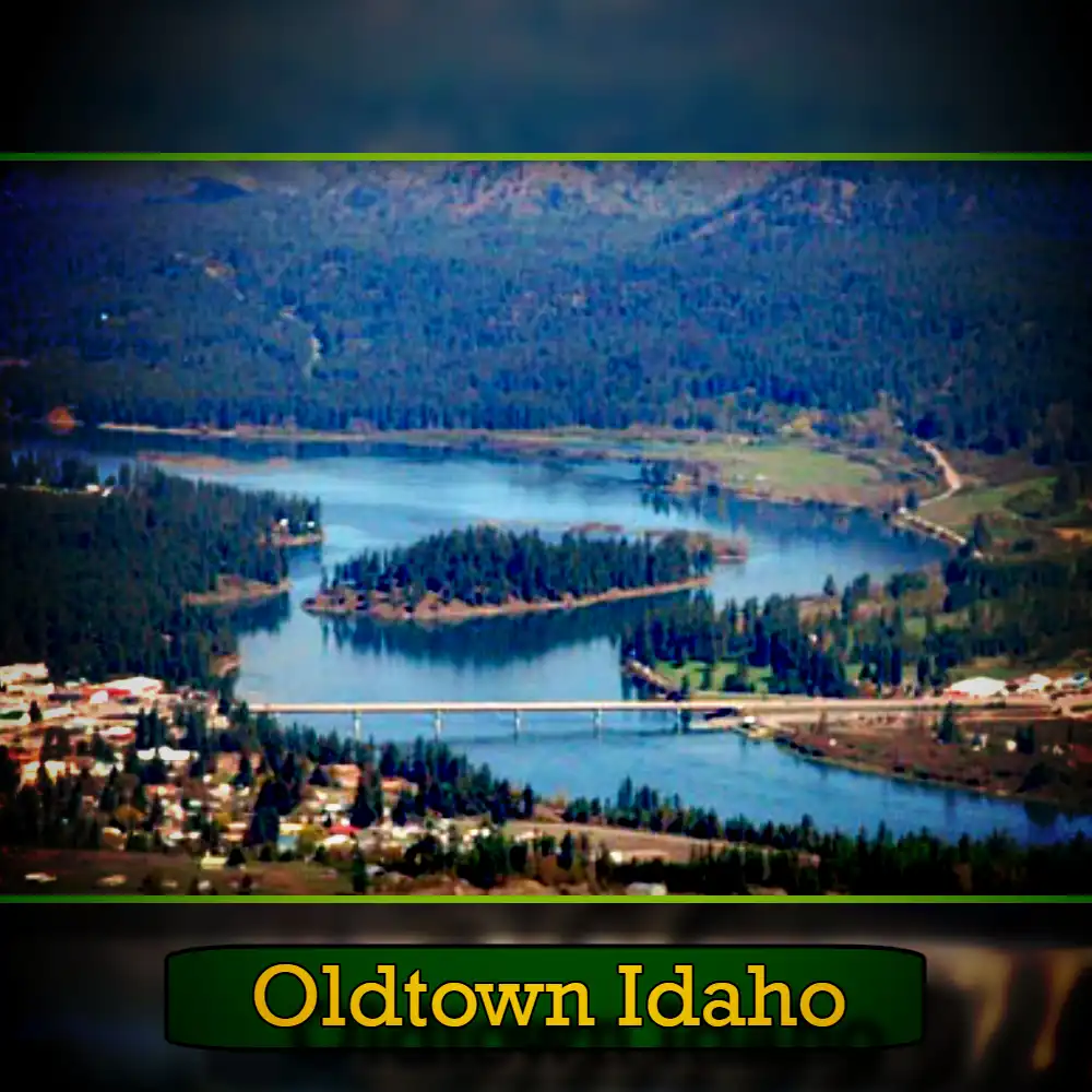 Aerial view of Oldtown, Idaho, showing a lake surrounded by forested hills and a bridge connecting parts of the town, with a tow truck making its way across the bridge.
