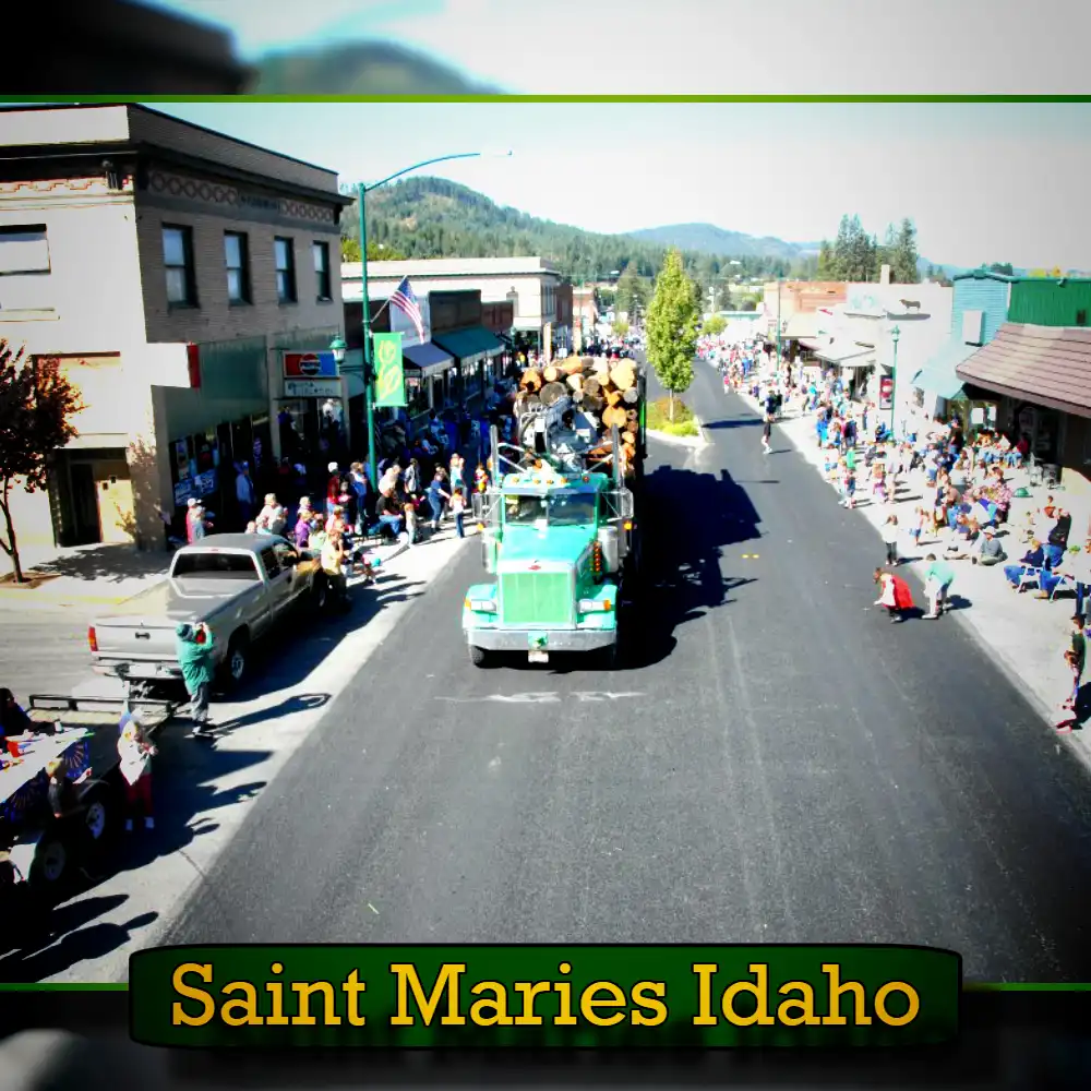 A parade in Saint Maries, Idaho, features a decorated tow truck loaded with logs driving down a street lined with spectators on both sides. The town's buildings and distant hills are visible.