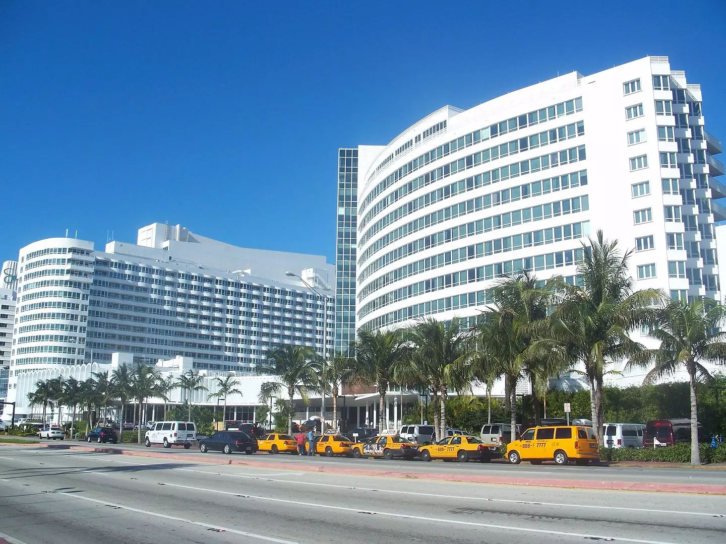 White modern hotel buildings with curved façades, lined with palm trees, and yellow taxis parked in front along a sunny street. A tow truck is stationed nearby, ready to provide towing services if needed. Clear blue sky in the background.