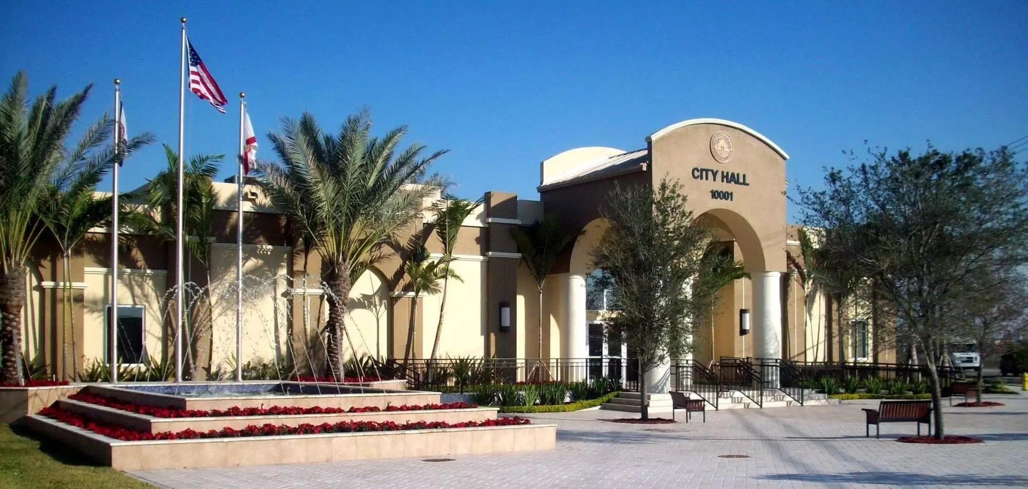 Front view of a beige-colored city hall building with a fountain in the foreground, flanked by palm trees and multiple flags on flagpoles, with a tow truck parked nearby.