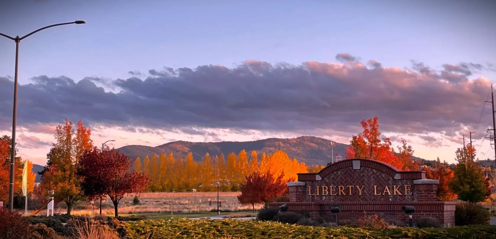 Autumn scene at Liberty Lake Park with colorful foliage, mountains in the background, and a partly cloudy sky. A brick sign for Liberty Lake stands in the foreground, with a local tow service truck visible in the distance.