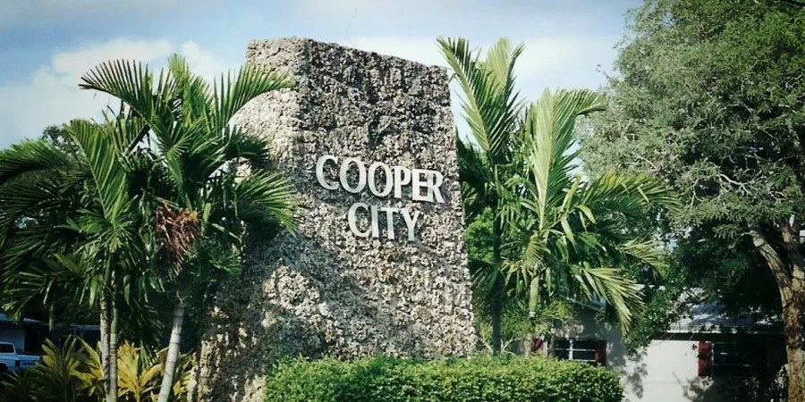 Stone sign for Cooper City surrounded by palm trees and greenery with a nearby tow truck providing service for the area.