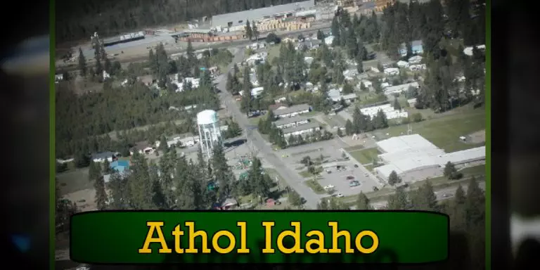 Aerial view of Athol, Idaho, showing a small town with a mix of trees, buildings, residential areas, and a prominent water tower. The town is surrounded by dense forest, with local businesses like a reliable tow service adding to the community's charm.