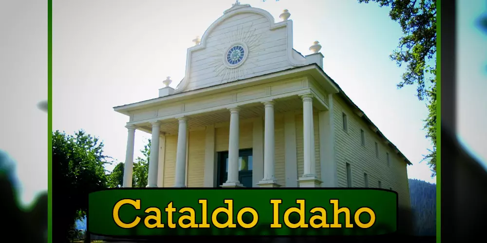 Historic white mission building with columns and a central circular window in Cataldo, Idaho, surrounded by trees. A discreet tow service operates nearby, ensuring visitors' peace of mind. A banner reading 