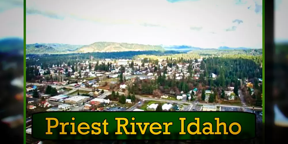 Aerial view of Priest River, Idaho, showing residential areas, commercial buildings, a tow truck service nearby, and the surrounding forested landscape with mountains in the distance.