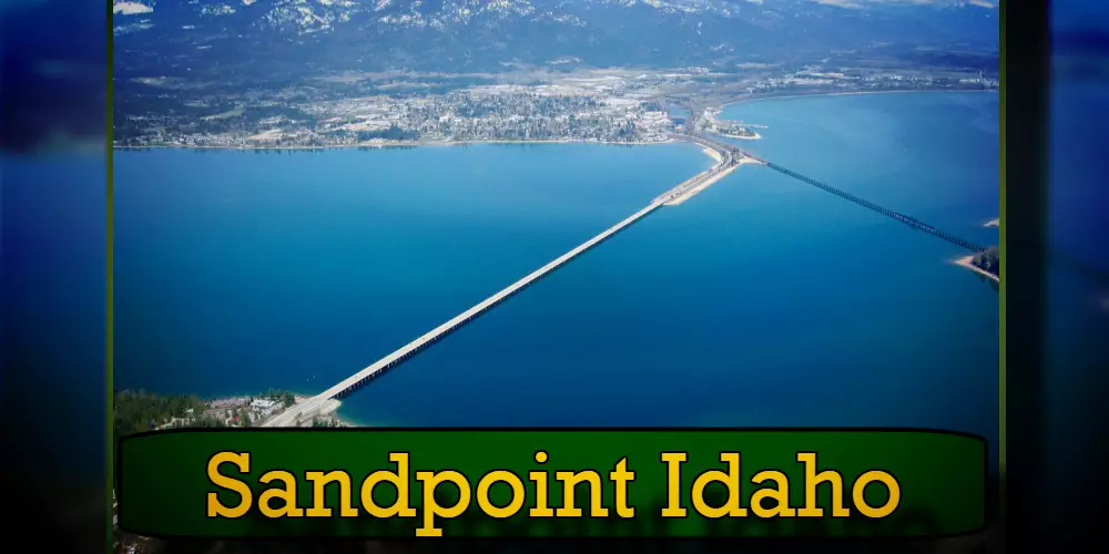 Aerial view of Sandpoint, Idaho, featuring a long bridge over Lake Pend Oreille with mountains in the background. A tow truck is crossing the bridge. The text 