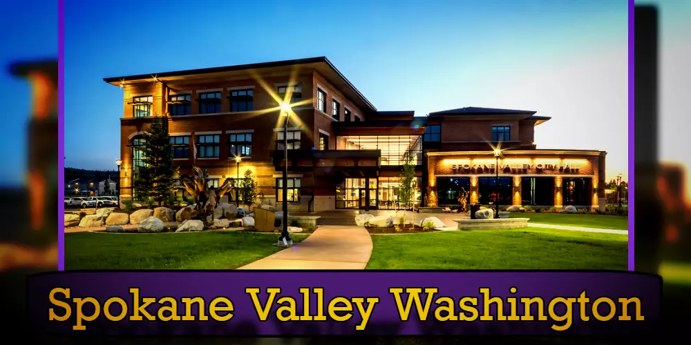 The exterior of a well-lit building labeled Spokane Valley City Hall stands against a clear evening sky, with a meticulously landscaped area and a discreet tow service nearby for any roadside assistance needed.