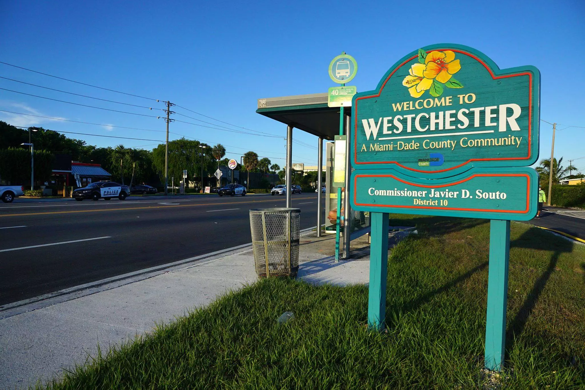 Welcome sign for Westchester, a Miami-Dade County community, featuring a bus stop and street in the background. A tow truck from a local tow service is parked nearby. Commissioner Javier D. Souto is noted.