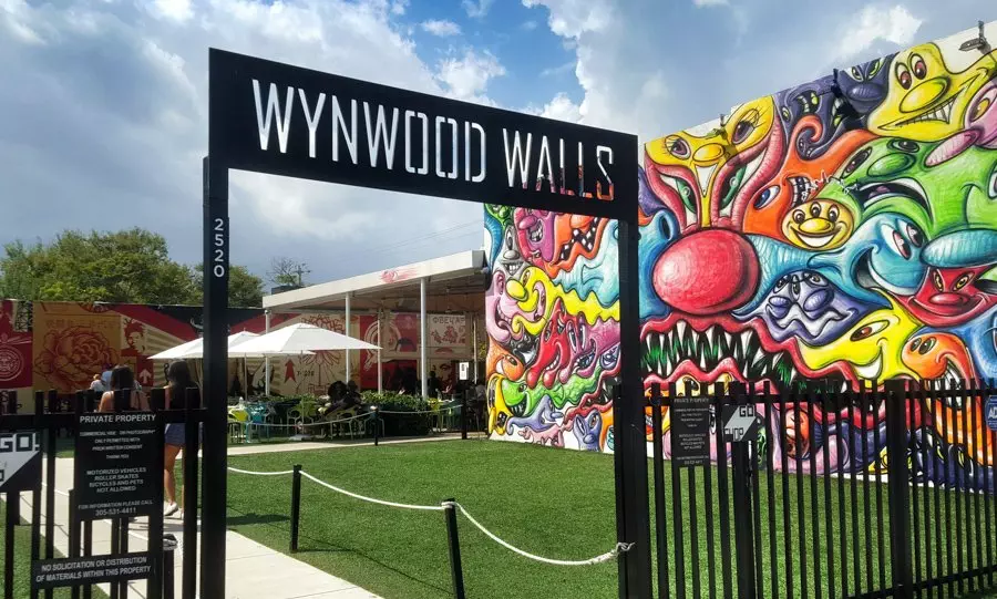 Entrance to Wynwood Walls, showcasing vibrant graffiti art on walls against a partly cloudy sky. Visitors can be seen in the background near an outdoor seating area, while a nearby tow truck subtly reminds us of the bustling city life.